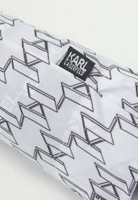 Biały woreczek z tkaniny z czarnymi wzorami geometrycznymi. Zawiera czarną metkę z napisem "KARL LAGERFELD" w srebrnym kolorze. Gładka faktura.