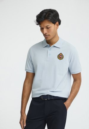 Uomo con polo azzurro chiaro ricamata con stemma e pantaloni scuri, in piedi con una mano in tasca, guarda verso il basso.