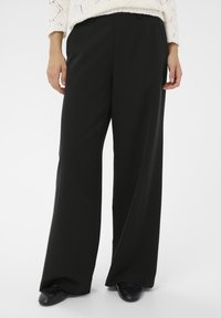 Fekete, széles lábú nadrág sima anyagból, testre szabott szabással és finom redőkkel, sötét slip-on cipővel párosítva.