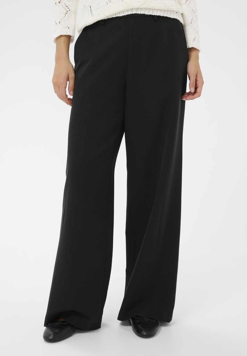 Fekete, széles lábú nadrág sima anyagból, testre szabott szabással és finom redőkkel, sötét slip-on cipővel párosítva.