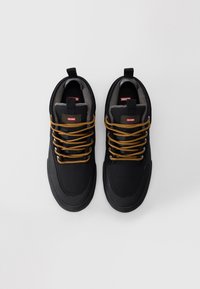 Pár černých tenisek Globe se žlutými tkaničkami, polstrovanými límci a poutky na světle šedém pozadí.