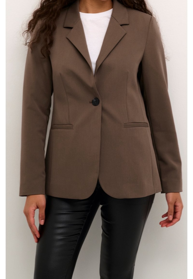 Femme portant un blazer marron ajusté sur une chemise blanche et un pantalon en cuir noir, debout devant un fond clair uni.