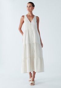 Kremowa maxi sukienka z dekoltem w kształcie V, z przodu zapinana na guziki, o warstwowej konstrukcji i miękkiej, teksturowanej tkaninie. Noszona z plecionymi sandałami.