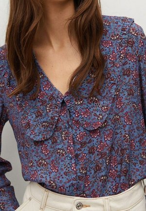 Vrouw draagt een blauwe blouse met lange mouwen en bloemenprint, voorzien van een grote gerimpelde kraag, ingestopt in een hoog taille lichtbeige broek.