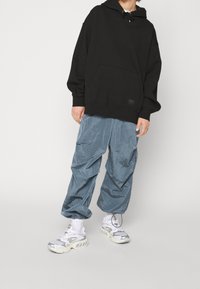 Persona vistiendo una sudadera con capucha negra de gran tamaño, pantalones cargo azules holgados, calcetines blancos y zapatillas gruesas plateado-blancas, de pie frente a un fondo liso.