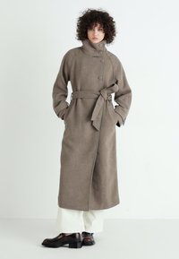 Casaco longo de lã taupe com gola alta, botões frontais, bolsos laterais e faixa de aperto. Textura suave, design minimalista.