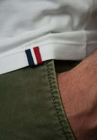 L'ourlet de la chemise blanche présente une rayure rouge, blanche et bleue. Un gros plan montre le tissu texturé vert du pantalon et une partie d'une main.
