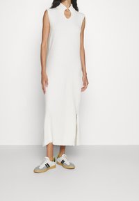 Abito in maglia bianco senza maniche con scollatura a chiave e spacco laterale, abbinato a sneakers grigie e bianche con dettagli neri e gialli.