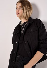 Czarna kurtka dżinsowa z dwoma kieszeniami na piersi, zapięciami na guziki i podwiniętymi rękawami; noszona na czarnym topie, ozdobiona warstwowymi srebrnymi naszyjnikami.