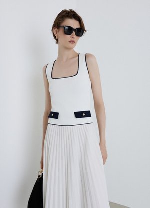 Frau mit schwarzen Sonnenbrillen, einem weißen ärmellosen Kleid mit schwarzem Besatz und plissiertem Rock, die eine schwarze Handtasche vor einem schlichten hellen Hintergrund hält.