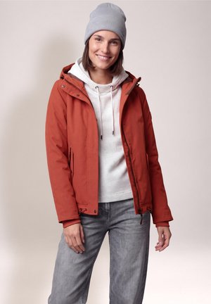 Femme souriante portant un bonnet gris, une veste couleur rouille, un sweat à capuche blanc et un jean gris, debout devant un fond clair uni.