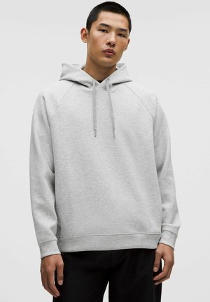 Junger Mann in einem hellgrauen Kapuzenpullover mit Kordelzügen, vor einem schlichten weißen Hintergrund stehend.