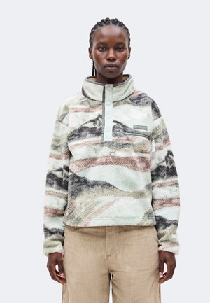 Persona que lleva un forro polar de cuello alto abotonado con patrones abstractos de montaña y pantalones de pana beige.