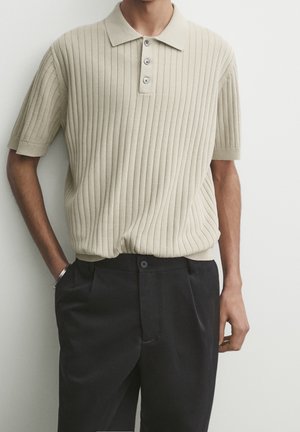 Homme portant un polo à manches courtes côtelé beige, rentré dans un pantalon noir taille haute, les mains dans les poches.