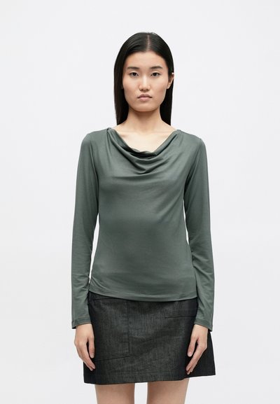 Top de manga longa em tecido verde suave e discreto com decote drapeado, combinado com uma saia cinza escura com bolsos laterais.