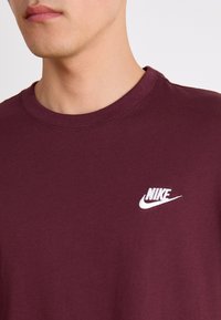 Läheltä otettu kuva henkilöstä, joka käyttää viininpunaista pyöreäkaulaista paitaa, jossa on valkoinen Nike-logo brodeerattuna vasempaan rintaan.