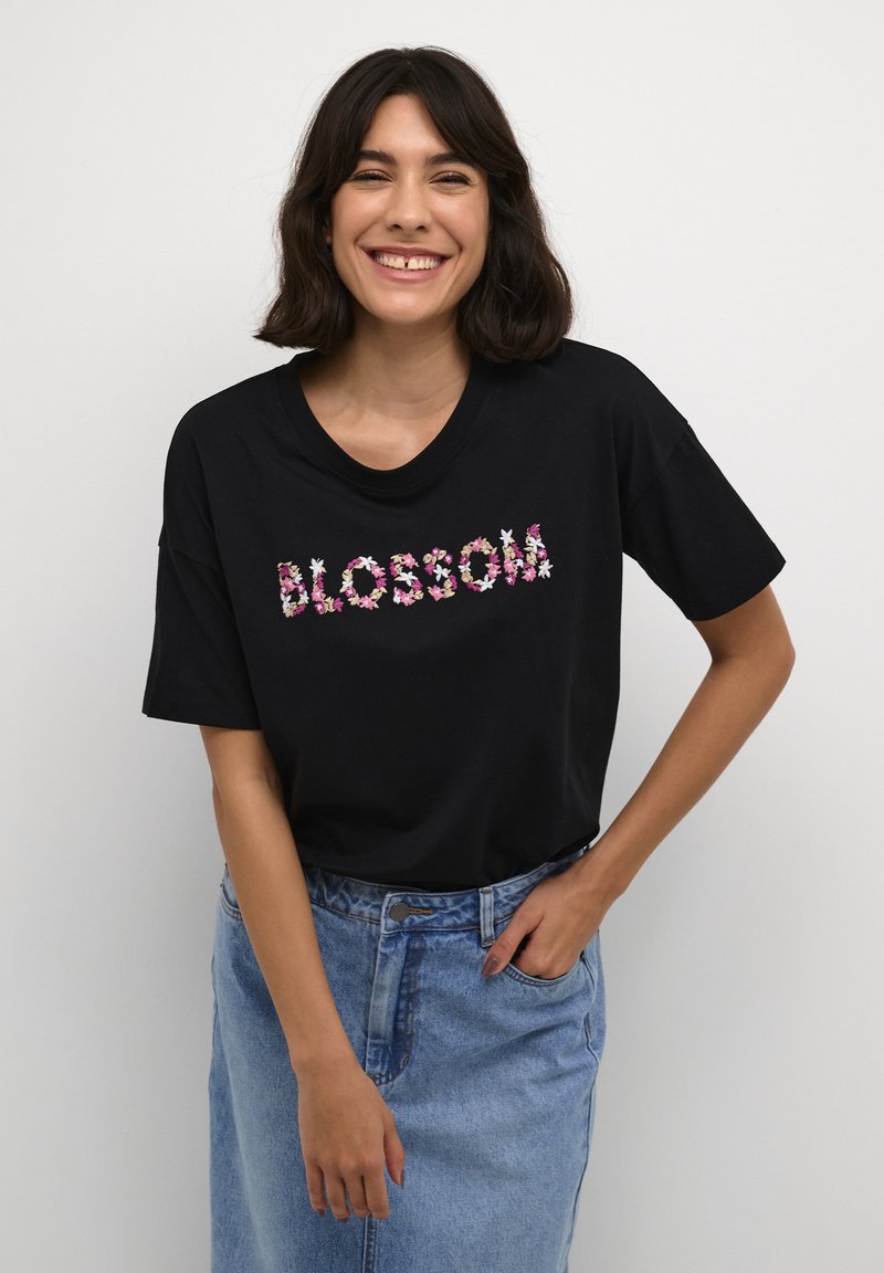 Crni pamučni majica s kratkim rukavima, s cvjetnim vezom koji ispisuje "BLOSSOM" u ružičastoj i bijeloj boji na jednobojnoj pozadini.