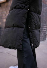 Manteau rembourré noir avec une texture matelassée, fermeture à boutons-pression et fente sur le côté, associé à un pantalon côtelé noir et des baskets blanches.