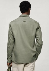 Camicia a maniche lunghe verde chiaro con una texture liscia, colletto a punta e polsini abbottonati. Presenta una piccola etichetta nella parte superiore della schiena.