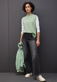 Jasnozielony sweter z dzianiny z białymi rękawami, czarne spodnie z szerokimi nogawkami i białe sneakersy. W jednej ręce trzymana jest pasująca zielona kurtka.