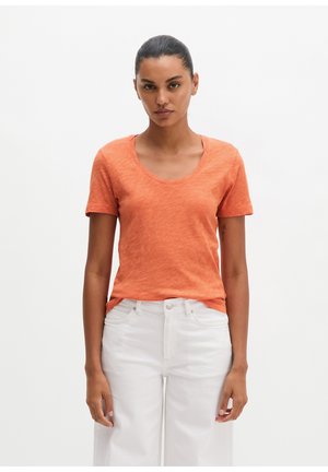 Femme aux cheveux attachés portant un t-shirt orange à manches courtes et col arrondi, et un pantalon blanc taille haute, debout devant un fond uni.