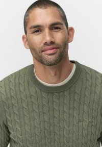 Grön kabelstickad tröja med rund hals, texturerad mönster, i kombination med en ljusfärgad innertröja. Ren, minimalistisk design.