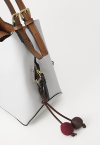Sac fourre-tout en cuir gris avec des anses en cuir marron, quincaillerie dorée et fermeture éclair. Présente un cordon décoratif avec deux pompons texturés en rouge et marron.
