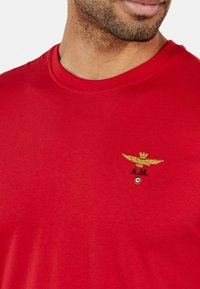 T-shirt rossa con scollatura a girocollo, con logo aquila ricamato in oro e testo "A.M." sul petto. Tessuto di cotone morbido.
