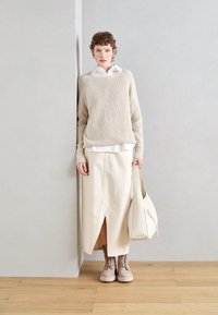 Beige ribbestrikket genser, hvit skjorte med krage under, kremfarget asymmetrisk skjørt med splitt, beige snøresko, og en hvit handlebag.