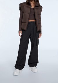 Bruine gewatteerde bodywarmer over een nauwsluitend donkerbruin shirt, gecombineerd met losse zwarte cargobroeken en witte chunky sneakers. Eenvoudig, functioneel ontwerp.