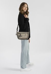 Graue Schultertasche mit verspielten Grafiken, silbernen Beschlägen und verstellbarem Tragegurt. Auf einem Modell mit schwarzem Blazer und hellen Jeans platziert.