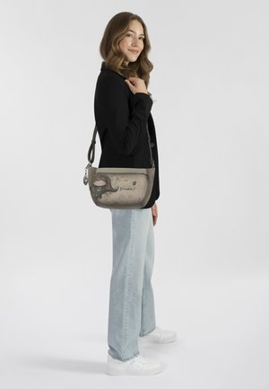Graue Schultertasche mit verspielten Grafiken, silbernen Beschlägen und verstellbarem Tragegurt. Auf einem Modell mit schwarzem Blazer und hellen Jeans platziert.