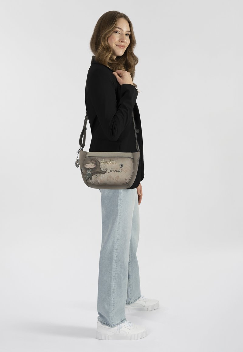 Graue Schultertasche mit verspielten Grafiken, silbernen Beschlägen und verstellbarem Tragegurt. Auf einem Modell mit schwarzem Blazer und hellen Jeans platziert.