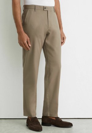 Mann trägt beige maßgeschneiderte Hose und dunkelbraune Wildleder-Loafers, steht vor einer weißen Wand.