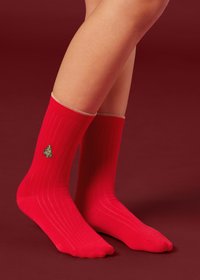 Calzini rossi a coste con un albero di Natale verde ricamato e un risvolto beige a contrasto, indossati su una gamba su uno sfondo rosso scuro.