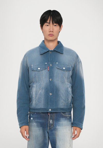 Young man wearing faded blue corduroy jacket with buttoned front and matching denim jeans with paint splatter and distressing details.