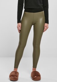 Persona che indossa leggings lucidi verde oliva, una maglietta nera e pantofole marroni pelose, in piedi contro uno sfondo grigio chiaro uniforme.