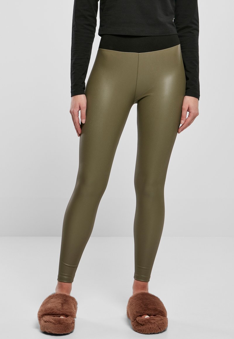 Persona che indossa leggings lucidi verde oliva, una maglietta nera e pantofole marroni pelose, in piedi contro uno sfondo grigio chiaro uniforme.