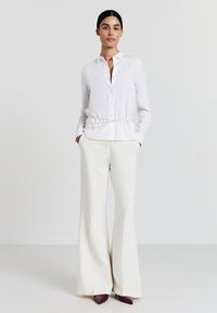Donna che indossa una camicia bianca con dettagli in catena di perle, pantaloni beige a gamba larga e scarpe viola con punta appuntita, in piedi di fronte a uno sfondo neutro.