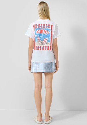 Femme aux longs cheveux blonds portant un t-shirt blanc avec un motif de parasol de plage rouge et blanc et de chaises, une jupe bleue rayée et des sandales blanches.