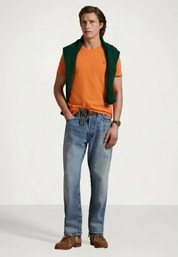 T-shirt laranja, camisola verde colocada sobre os ombros, jeans azuis, sapatos castanhos, design simples, ajuste casual, sem padrões visíveis.