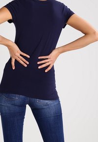 Henkilö, jolla on yllään tummansininen paita ja siniset farkut, pitää molemmilla käsillään alaselkäänsä, mikä viittaa selkäkipuun tai epämukavuuteen.