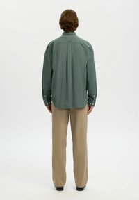 Homme debout, de dos, portant une chemise verte ample à boutons, un pantalon beige et des chaussures noires sur un fond blanc.