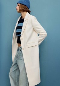 Cappotto lungo color crema con un design su misura, caratterizzato da due bottoni e tasche laterali. Indossato sopra un maglione corto a righe blu e nero e jeans azzurri.