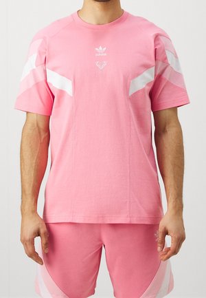 Mann trägt ein pinkes Adidas T-Shirt mit kurzen Ärmeln und passenden Shorts mit weißen Chevron-Streifen an Ärmeln und Beinen.