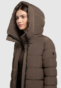 Giacca imbottita marrone con un grande cappuccio strutturato. Presenta una chiusura con zip e bottoni a pressione, una texture liscia e un dettaglio con logo sul maniche.