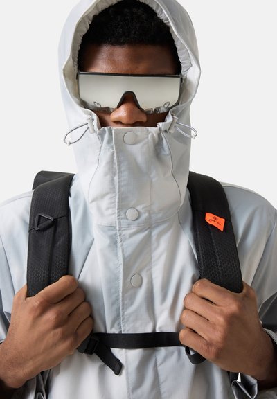 Osoba w jasnoszarej kurtce z kapturem, zapiętym pod brodą, nosząca refleksyjne gogle narciarskie i czarne paski plecaka.