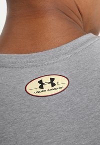 Harmaa kangaspaita, jossa on lyhyet hihat, ja joka on varustettu pyöreällä tarralapulla, jossa on musta logo ja teksti "UNDER ARMOUR" takakäyrässä.