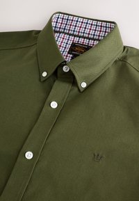 Olivovo zelená košeľa na gombíky s bielymi gombíkmi, kockovaným vnútorným lemom goliera a malým vyšívaným logom koruny na hrudníku.