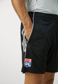 Svarte idrettsshorts laget av glatt stoff, med hvite sidestriper og et brodert Olympique Lyonnais-logo på venstre ben.